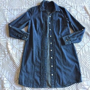 May include: A long-sleeved denim dress in a medium blue wash. The dress features a button-down front with white buttons and a ruffled trim down the center. The dress has a collared neckline and buttoned cuffs.