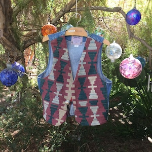 K&ouml;nnte beinhalten: Eine Jeansweste mit einem bunten geometrischen Muster auf der Vorderseite. Die Weste h&auml;ngt an einem Holzb&uuml;gel und ist von Glasornamenten umgeben.