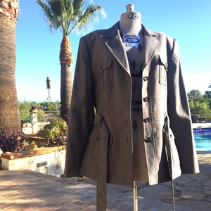 May include: A gray tweed blazer with a belt and flap pockets. The blazer has a three-button closure and a notched lapel.
