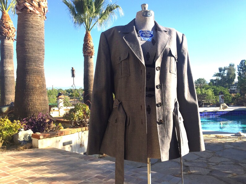 May include: A gray tweed blazer with a belt and flap pockets. The blazer has a three-button closure and a notched lapel.