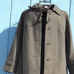 May include: A long, olive-green coat with a collared neckline and button-down closure. The coat features a classic design with a tailored fit, visible buttons, and a structured silhouette. The coat is displayed against a light blue wooden background.