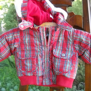 May include: A red, green, and blue plaid baby jacket with a red hood lined with white faux fur. The jacket has a zipper closure, two front pockets, and a red ribbed hem and cuffs. The jacket is hanging on a wooden hanger.
