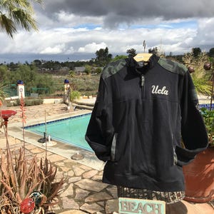 Puede incluir: Sudadera negra y verde camuflaje con cremallera de 1/4 y la palabra "UCLA" bordada en blanco en la parte delantera.