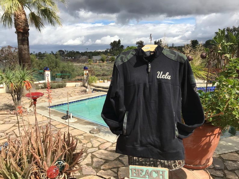 Puede incluir: Sudadera negra y verde camuflaje con cremallera de 1/4 y la palabra "UCLA" bordada en blanco en la parte delantera.