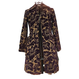 May include: A long-sleeved coat featuring a dark purple floral pattern on a tan background. The coat has a structured collar, a button-front closure, and two front pockets with flaps. The fabric has a textured appearance with a repeating botanical design.