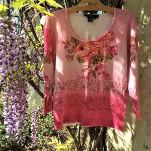 May include: A pink long-sleeve sweater with a scoop neck, featuring a floral design with sequins and bead embellishments. The sweater has a gradient color scheme, transitioning from light pink to a darker shade at the hem. The floral design includes roses and leaves.