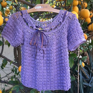 May include: A purple crocheted top with short sleeves and a lace neckline. The top has a ribbon tied at the neckline.