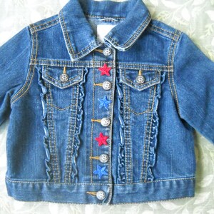 May include: A blue denim jacket with a classic collar and long sleeves. The jacket features decorative silver buttons and red and blue star appliqu&eacute;s. Ruffled trim accents the front, adding a touch of detail to this children's garment.