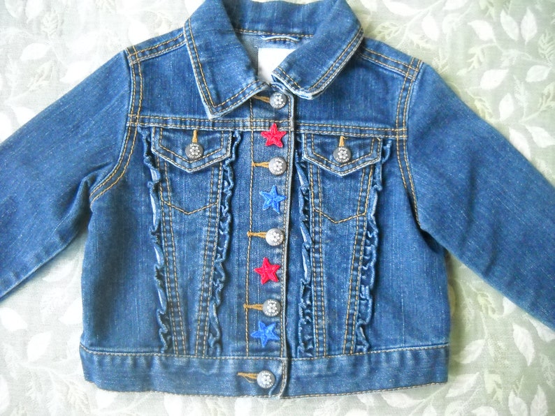 May include: A blue denim jacket with a classic collar and long sleeves. The jacket features decorative silver buttons and red and blue star appliqu&eacute;s. Ruffled trim accents the front, adding a touch of detail to this children's garment.