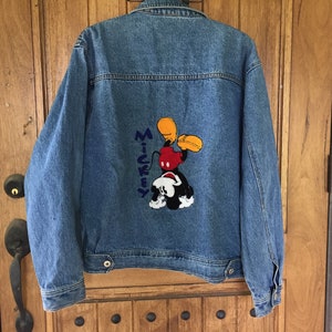 May include: A blue denim jacket with a Mickey Mouse applique on the back. The applique is yellow, red, black, and white. The text "Mickey" is embroidered in blue on the jacket.