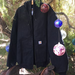 May include: A black Carhartt hooded jacket with a zipper closure and a small Carhartt logo patch on the left chest. The jacket is hanging on a tree branch with colorful glass ornaments hanging from the branch.