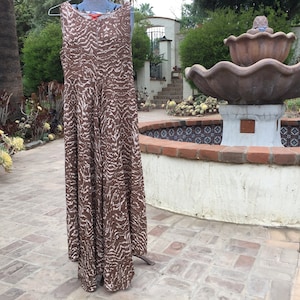 May include: A sleeveless, floor-length dress with a brown and white zebra print pattern. The dress is hanging on a hanger outdoors, with a fountain and greenery in the background. The dress has a flowing, A-line silhouette.