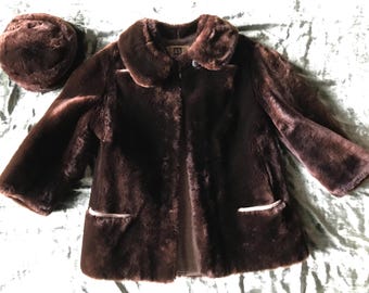 May include: A brown faux fur jacket with a matching hat. The jacket has a collar, long sleeves, and two pockets. The hat is a cloche style.