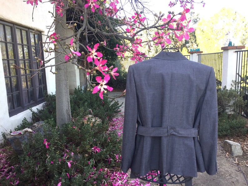 May include: A gray blazer with a belt, photographed against a backdrop of a tree with pink blossoms.