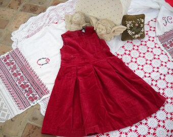 May include: A red corduroy dress with a white button and a white lace trim. The dress is on a white tablecloth with a red and white embroidered design.
