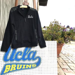 Puede incluir: Sudadera negra de cremallera de un cuarto con un patr&oacute;n de camuflaje en los hombros y un logotipo "UCLA" blanco en el pecho.