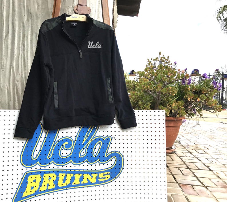 Puede incluir: Sudadera negra de cremallera de un cuarto con un patr&oacute;n de camuflaje en los hombros y un logotipo "UCLA" blanco en el pecho.
