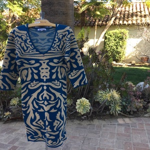 May include: A blue and beige patterned sweater dress with a V-neckline. The dress is hanging on a wooden hanger.
