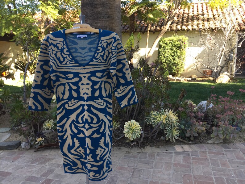 May include: A blue and beige patterned sweater dress with a V-neckline. The dress is hanging on a wooden hanger.