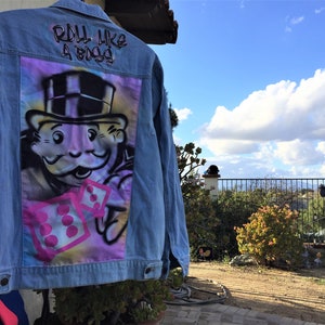 May include: A blue denim jacket with a colorful spray-painted design on the back. The design features a cartoon character wearing a top hat and holding dice. The text "Roll Like A Boss" is written in black above the design.