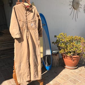 May include: A tan work jumpsuit with a button-up front, collar, and multiple pockets. The jumpsuit is hanging on a wooden hanger. A blue and white surfboard leans against a wall in the background.