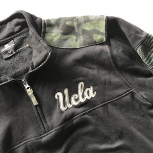 Puede incluir: Sudadera UCLA negra con un patr&oacute;n de camuflaje en el hombro y un peque&ntilde;o parche de la bandera estadounidense en la manga. La sudadera tiene una cremallera y la palabra "UCLA" est&aacute; bordada en la parte delantera.