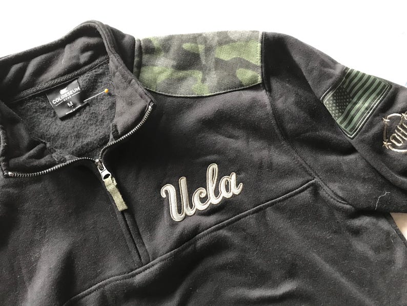 Puede incluir: Sudadera UCLA negra con un patr&oacute;n de camuflaje en el hombro y un peque&ntilde;o parche de la bandera estadounidense en la manga. La sudadera tiene una cremallera y la palabra "UCLA" est&aacute; bordada en la parte delantera.