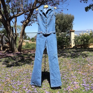 May include: A blue denim jumpsuit with a collared sleeveless top, a button-up front, and a belt. The jumpsuit has a wide-leg style.