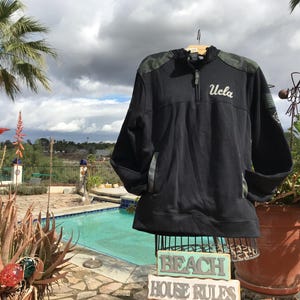 Puede incluir: Sudadera negra y verde de camuflaje con cremallera y un logotipo blanco "UCLA" en la parte delantera. La sudadera est&aacute; en un maniqu&iacute; delante de una piscina.
