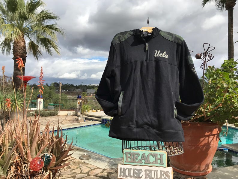 Puede incluir: Sudadera negra y verde de camuflaje con cremallera y un logotipo blanco "UCLA" en la parte delantera. La sudadera est&aacute; en un maniqu&iacute; delante de una piscina.