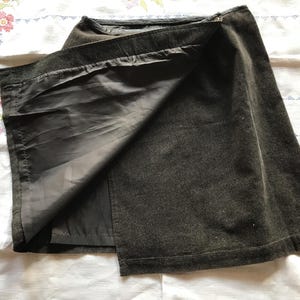 May include: A dark charcoal gray wrap skirt featuring a straight hemline and a smooth, solid-colored lining visible on the interior. The exterior fabric has a subtle, textured appearance, and the garment is constructed with a clean, simple design.