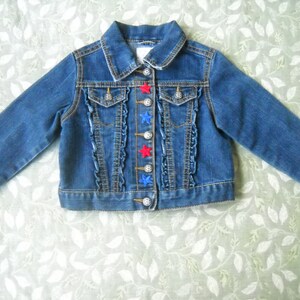 May include: A blue denim jacket with long sleeves. The jacket has a classic collar, two chest pockets, and decorative ruffles down the front. Red and blue star-shaped buttons add a playful touch to the design.