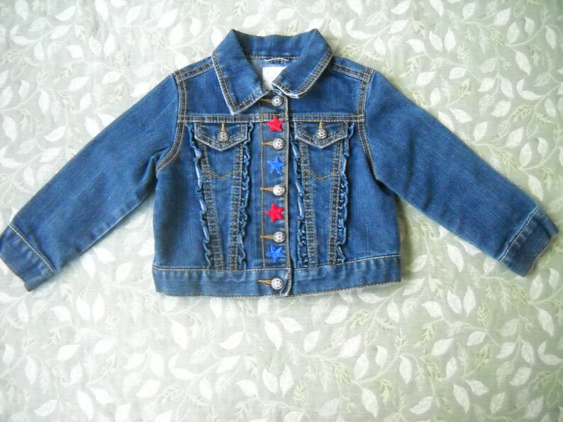 May include: A blue denim jacket with long sleeves. The jacket has a classic collar, two chest pockets, and decorative ruffles down the front. Red and blue star-shaped buttons add a playful touch to the design.