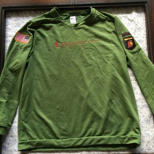 May include: Green long-sleeved sweatshirt with a red, white and blue American flag patch on the left sleeve and a red, yellow and black patch with a helmet on the right sleeve. The sweatshirt has the text "The Army of the United States of America" in red on the front.