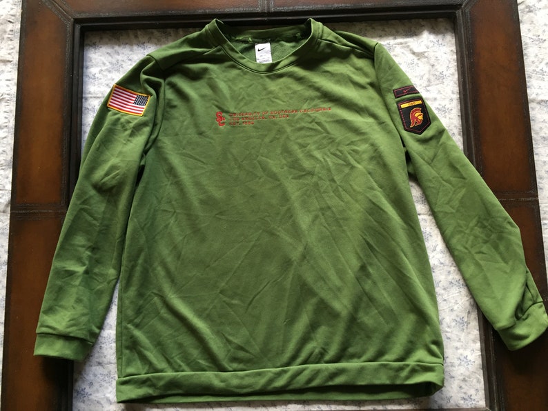 May include: Green long-sleeved sweatshirt with a red, white and blue American flag patch on the left sleeve and a red, yellow and black patch with a helmet on the right sleeve. The sweatshirt has the text "The Army of the United States of America" in red on the front.