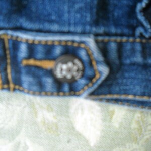 May include: Close-up of a section of a blue denim jacket. The jacket features a button with a logo and is stitched with yellow thread. The fabric has a worn appearance.
