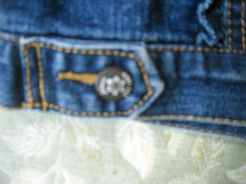 May include: Close-up of a section of a blue denim jacket. The jacket features a button with a logo and is stitched with yellow thread. The fabric has a worn appearance.