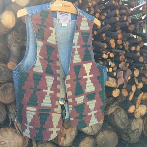 K&ouml;nnte beinhalten: Eine Jeansweste mit einem farbenfrohen geometrischen Muster auf der Vorderseite. Die Weste hat einen Knopfverschluss und eine kleine Tasche auf der linken Seite. Die Weste h&auml;ngt an einem Holzb&uuml;gel.