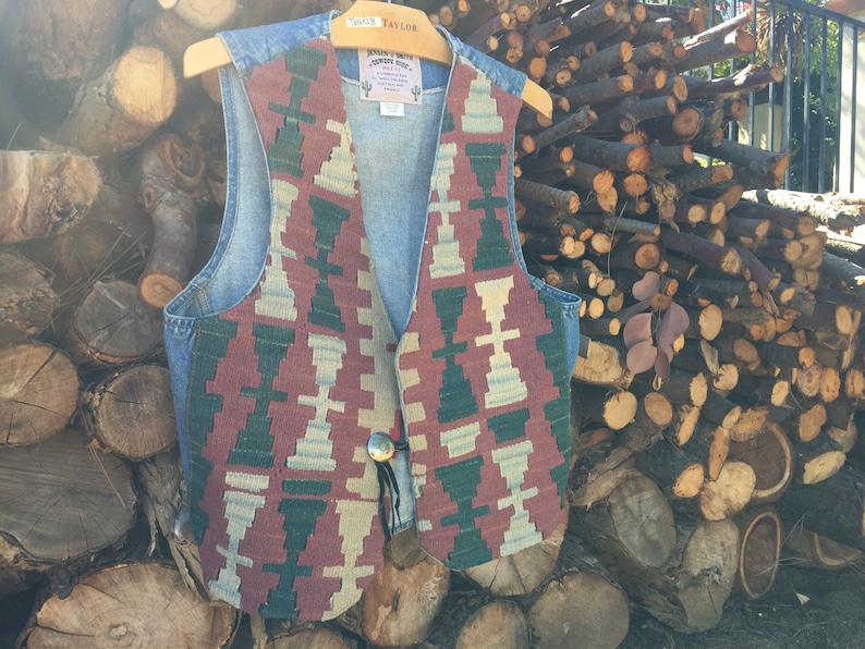 K&ouml;nnte beinhalten: Eine Jeansweste mit einem farbenfrohen geometrischen Muster auf der Vorderseite. Die Weste hat einen Knopfverschluss und eine kleine Tasche auf der linken Seite. Die Weste h&auml;ngt an einem Holzb&uuml;gel.