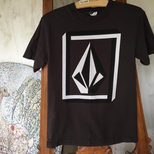 Pu&ograve; includere: Una maglietta nera con una grafica bianca a forma di diamante all'interno di un quadrato. La forma del diamante &egrave; delineata in nero e il quadrato &egrave; delineato in bianco.