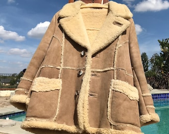 May include: A brown suede coat with a cream-colored shearling lining and trim. The coat features large pockets, button closures, and a wide collar. The coat is displayed on a wooden easel against a blue sky background.