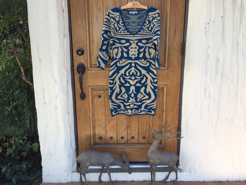 May include: A teal and beige patterned dress with a v-neck and 3/4 sleeves hanging on a hanger in front of a wooden door.