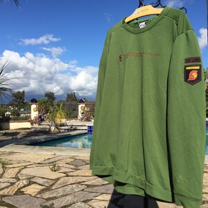 May include: Green long-sleeved sweatshirt with a Nike logo and a patch featuring a Spartan helmet on the right sleeve. The text "UNIVERSITY OF SOUTHERN CALIFORNIA" is printed on the front of the sweatshirt.