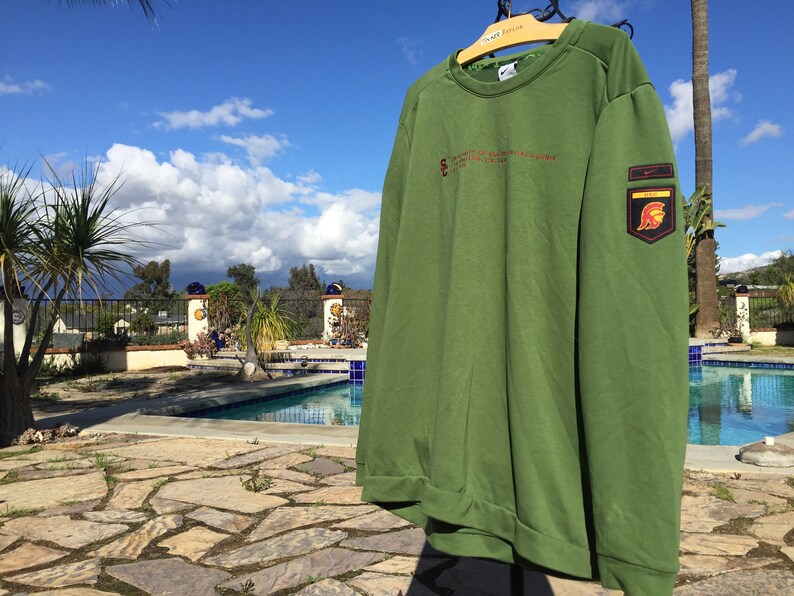 May include: Green long-sleeved sweatshirt with a Nike logo and a patch featuring a Spartan helmet on the right sleeve. The text "UNIVERSITY OF SOUTHERN CALIFORNIA" is printed on the front of the sweatshirt.