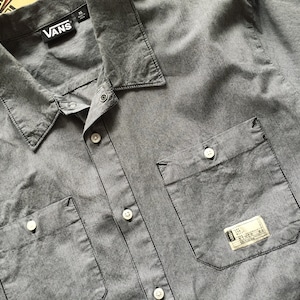 May include: Gray button-up shirt with two chest pockets and a collar. The shirt has the "Vans" logo on the collar and a small tag on the pocket. The shirt is XL size.