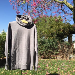 May include: A gray hooded sweatshirt with a pouch pocket. The sweatshirt is hanging on a hanger in front of a green bush and a tree with pink flowers.