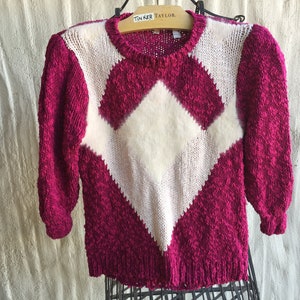 May include: A pink and white knit sweater with a diamond pattern. The sweater has a crew neck and long sleeves.
