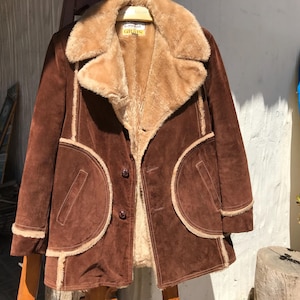 May include: A brown suede coat with a tan faux fur collar and lining. The coat features large, rounded pockets and trim in a lighter tan color. It has three buttons and is hanging on a wooden hanger.