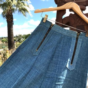 May include: A high-waisted blue denim skirt featuring two vertical dark metal zippers on the front. The skirt has a structured waistband and is suspended from a light-colored wooden hanger, showcasing a textured fabric finish and clean lines.