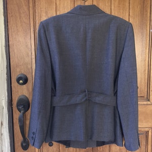 May include: A dark gray blazer with a belt in the back. The blazer has a classic style and is made of a textured fabric.